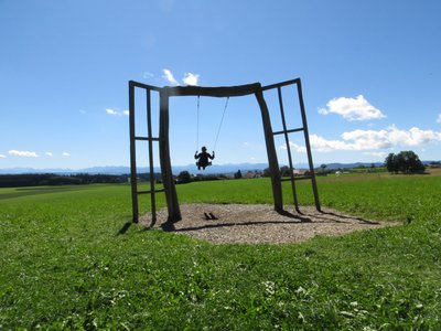 Große Holzschaukel auf Wiese; Person schaukelt vor dem allgäuer Bergpanorama